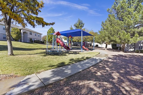 A playground with a red slide and a blue canopy is surrounded by a gravel path and trees.