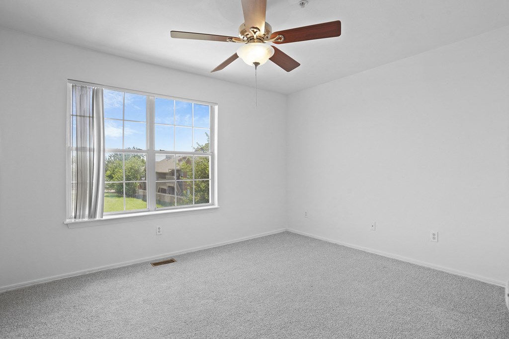 A room with a ceiling fan and a window overlooking a green area.