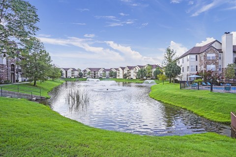 A serene lake with a fountain in the middle of a grassy area with houses in the background.