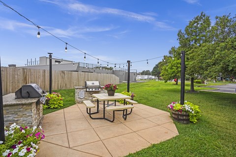 A patio with a picnic table and chairs is surrounded by a fence and has string lights above it.