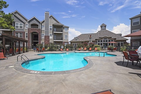 A large outdoor swimming pool surrounded by a patio with chairs and a building in the background.