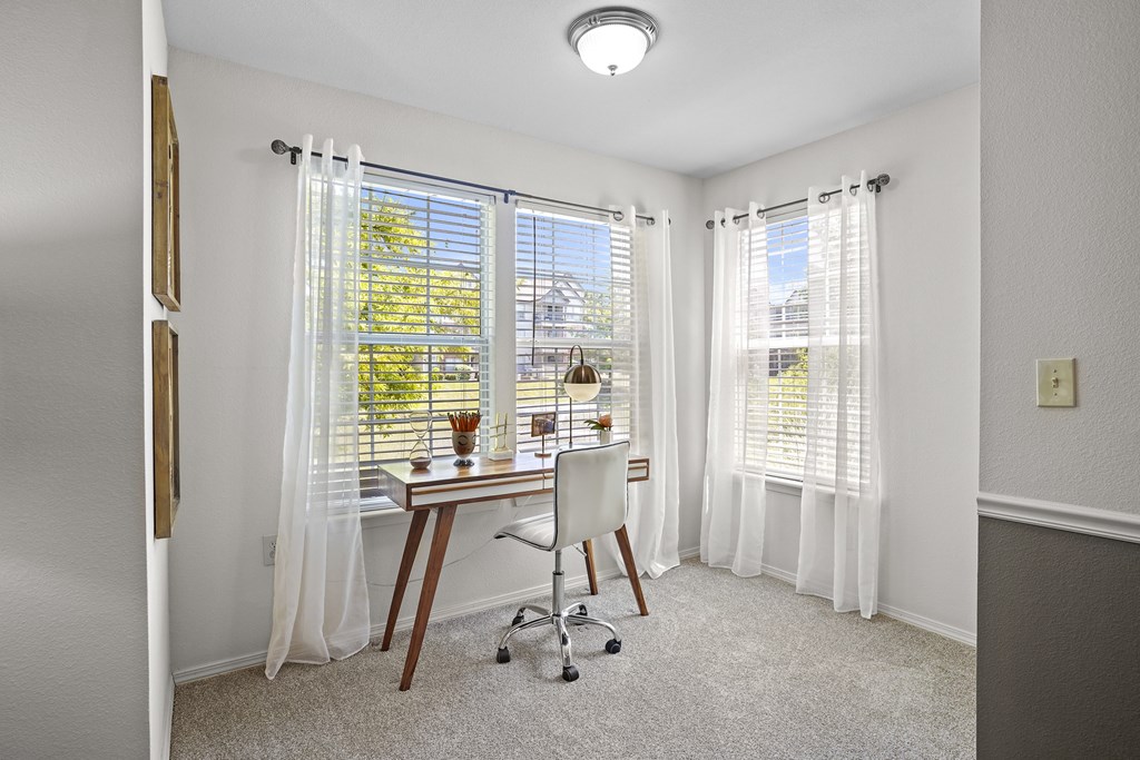 A room with a desk and chair in front of a window with white curtains.