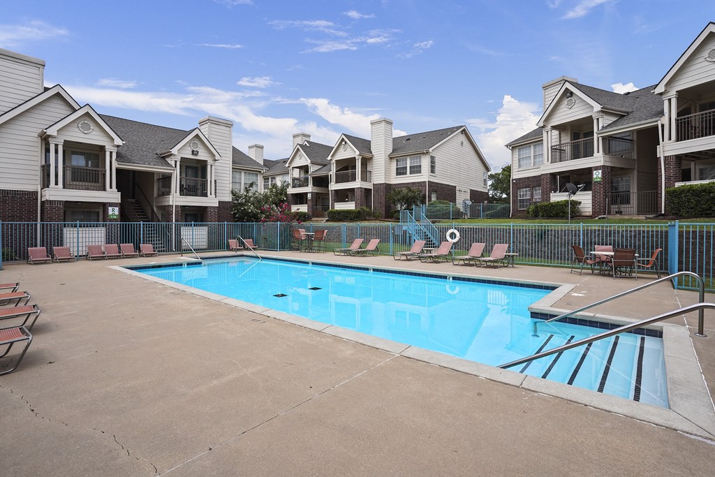 A swimming pool surrounded by a fence and chairs in front of apartment buildings.