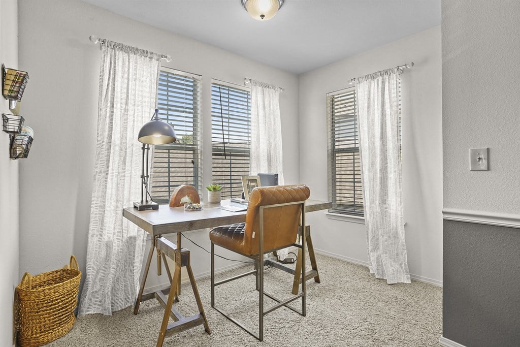 A room with a desk and chair in front of a window with white curtains.