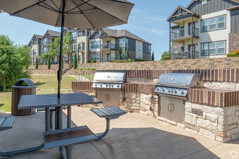 A patio with a table, chairs and an umbrella is set up in front of a stone wall and apartment buildings.