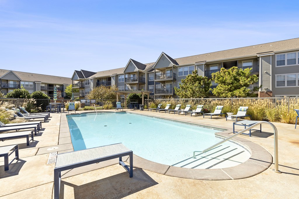 A swimming pool surrounded by sun loungers and chairs in front of apartment buildings.