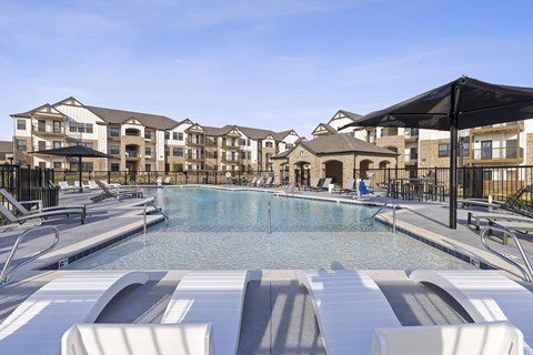 A large swimming pool surrounded by lounge chairs and umbrellas in front of apartment buildings.