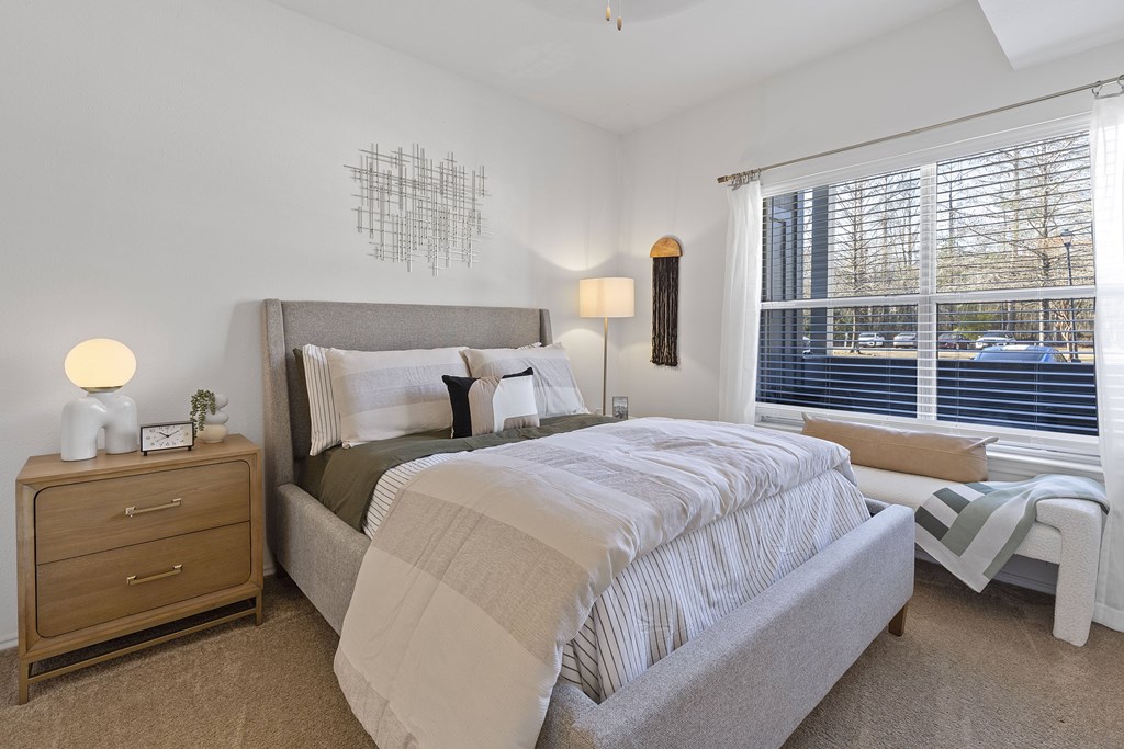 A bedroom with a large bed, a nightstand with a lamp, and a window with blinds.
