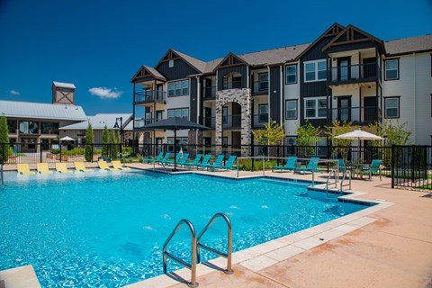 A large swimming pool surrounded by lounge chairs and umbrellas in front of a multi-story apartment building.