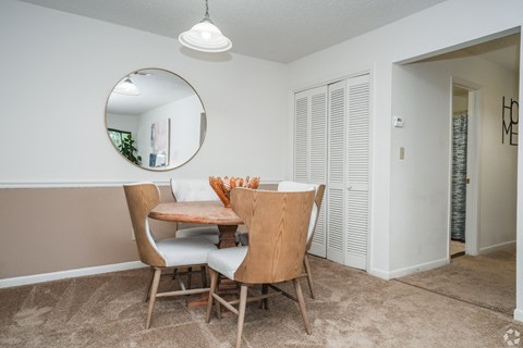 A dining room with a round mirror, wooden chairs, and a table with a white tablecloth and a vase of flowers.