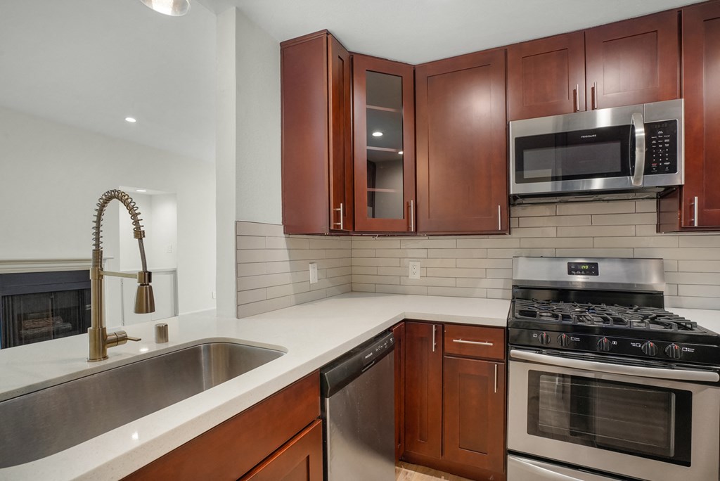 Apartment kitchen with stainless steel appliances, white tile back splash, white counter tops, and dark wood cabinets.