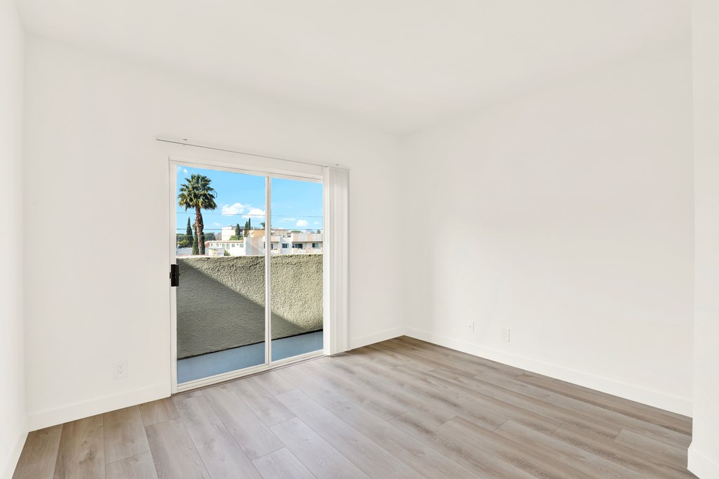 A room with a wooden floor and a window that shows a view of a palm tree and buildings outside.