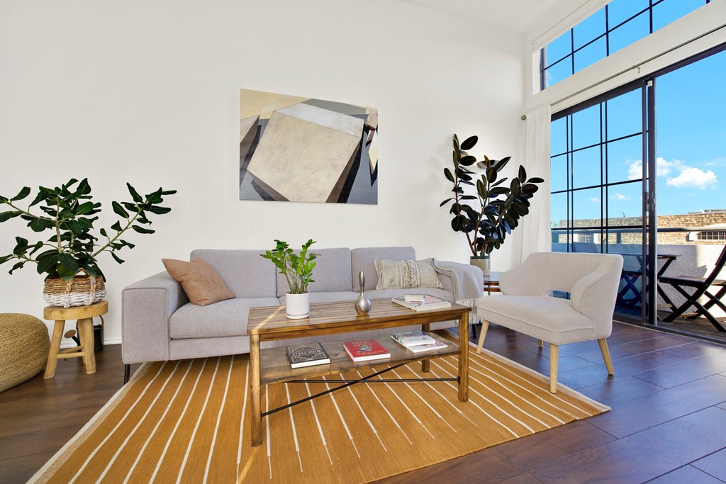 A living room with a grey couch, a white chair, a coffee table, and a large window at Beverly Wooster Apartments, Los Angeles