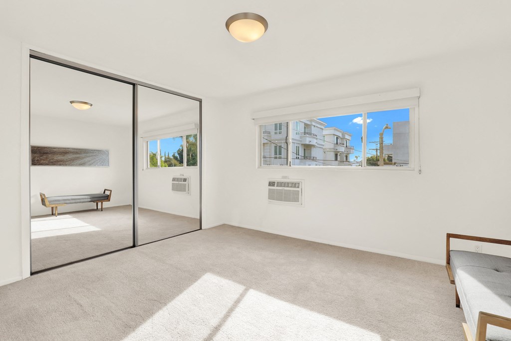 A white room with a bed and a window showing a view of a house at Wooster Encore Apartments, Los Angeles, California