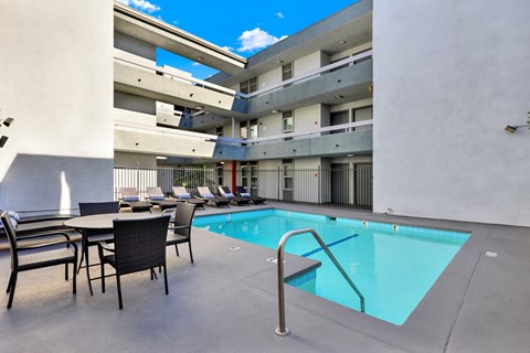 A pool area with chairs and a table is surrounded by apartment buildings.