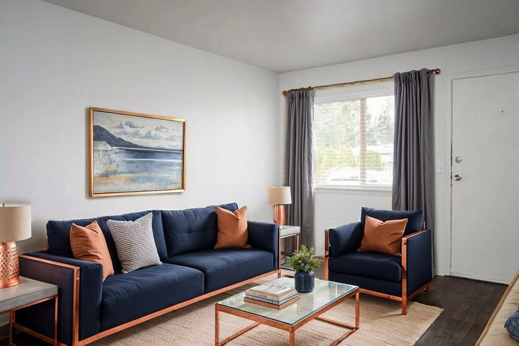 A living room with a blue sofa, a painting on the wall, and a glass table with a vase on it.