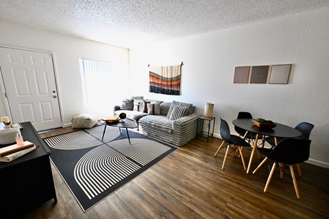 A living room with a black and white rug, a grey sofa, a black dining table and chairs, and a wooden floor.