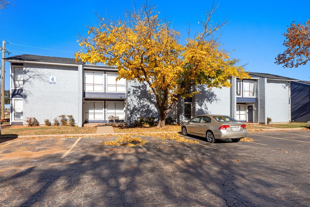 A silver car is parked in front of a grey building with a tree in front of it.