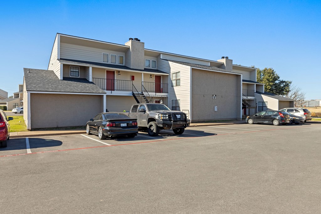 A black car is parked in a parking lot in front of a two-story apartment building.