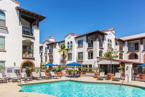A large swimming pool is surrounded by lounge chairs and umbrellas in front of a row of apartment buildings.
