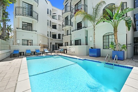 A large swimming pool in front of a white building with blue chairs around it.