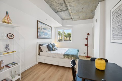 A new studio bedroom with a bed, a table, and a chair.  Palm trees are in the window.