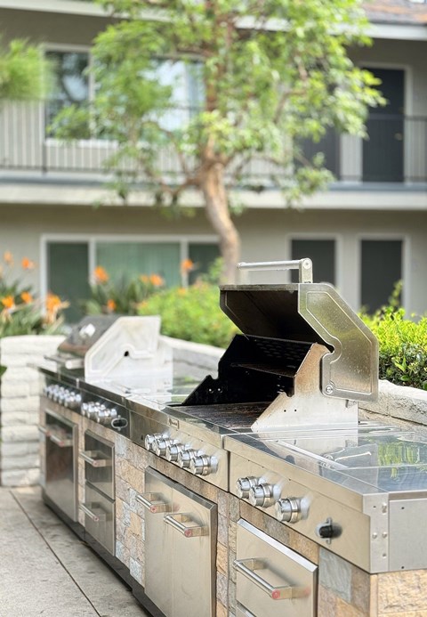 A BBQ grill with a clear lid is in front of a building with a tree and flowers in the background.