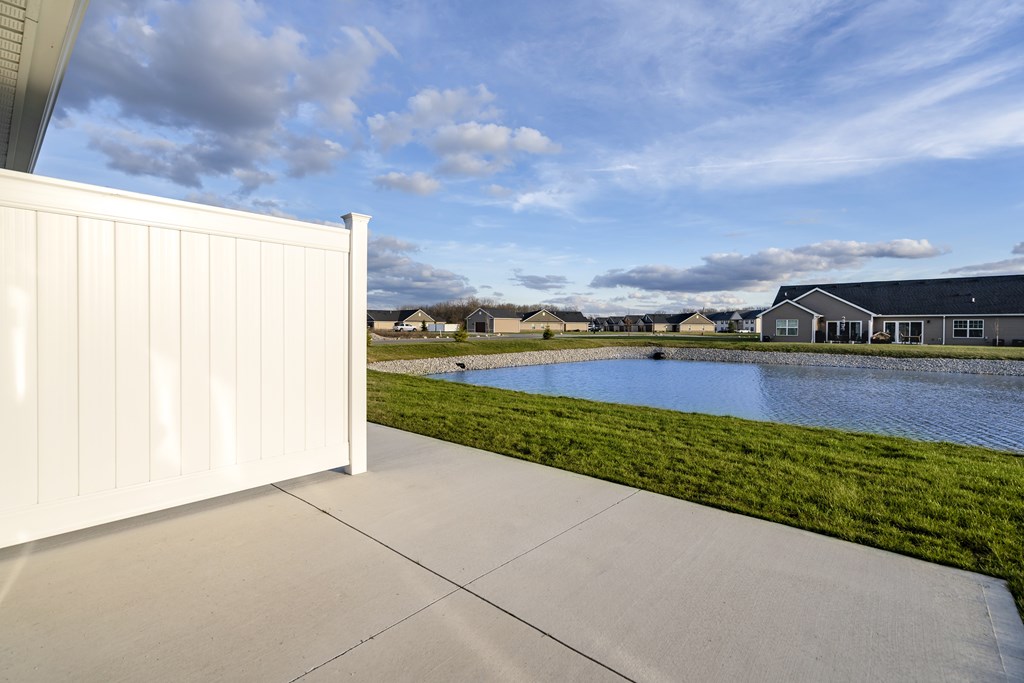 A white fence is in the foreground of a bright day with a house and a pond in the background.