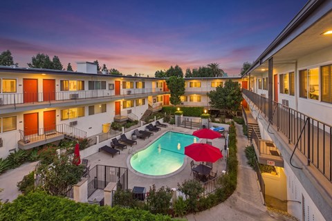 A pool surrounded by lounge chairs and umbrellas is the centerpiece of a courtyard surrounded by apartment buildings.