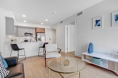 A modern living room with a glass coffee table and a kitchen area in the background.