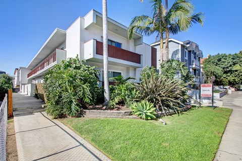 A white building with a red balcony is surrounded by greenery.