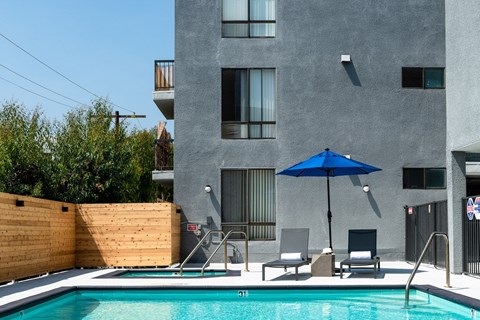 a pool with a blue umbrella and two chairs in front of a gray building