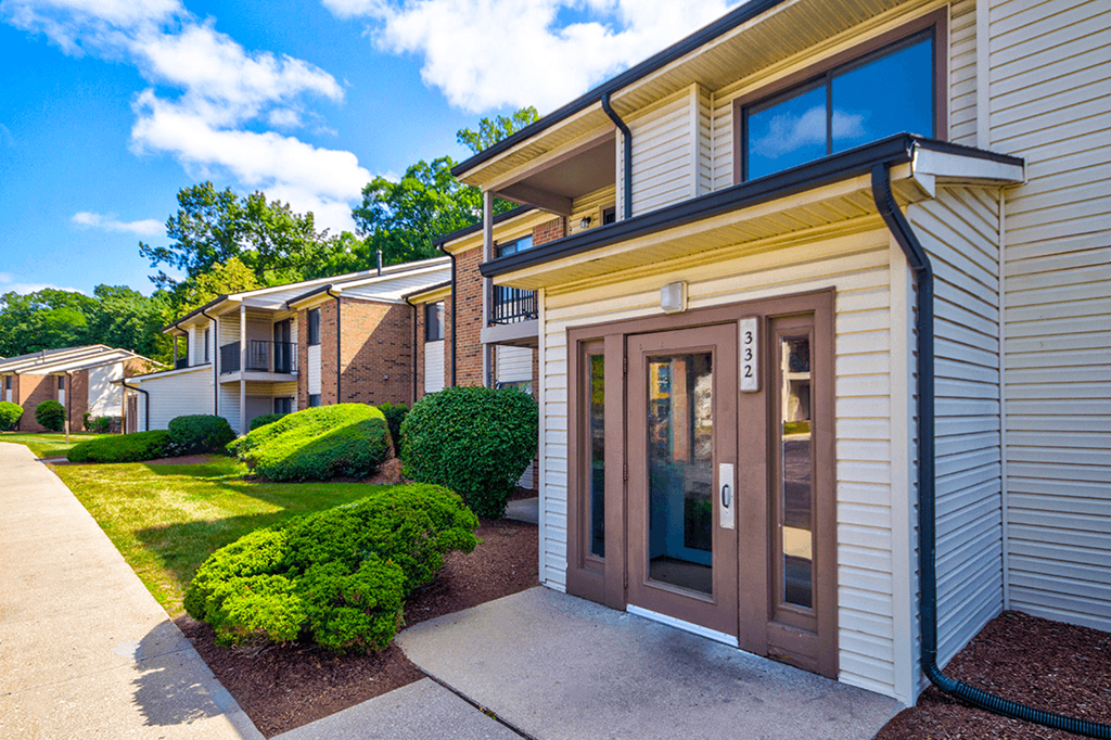 Exterior view of an apartment building at Woodland Crossing, showcasing landscaped surroundings, a sidewalk in front of the building, and a door leading into the entrance.