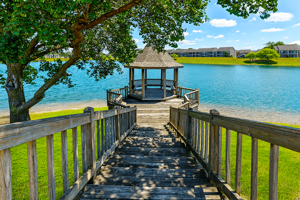A wooden walkway leads to a gazebo by a lake at Somerset Lakes Apartments, offering a tranquil outdoor retreat.