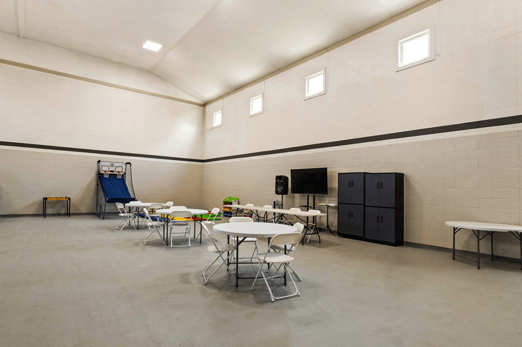 Indoor gymnasium at Windsor Park Apartments, featuring arcade basket ball equipment, a large area with tables and chairs and bright lighting.