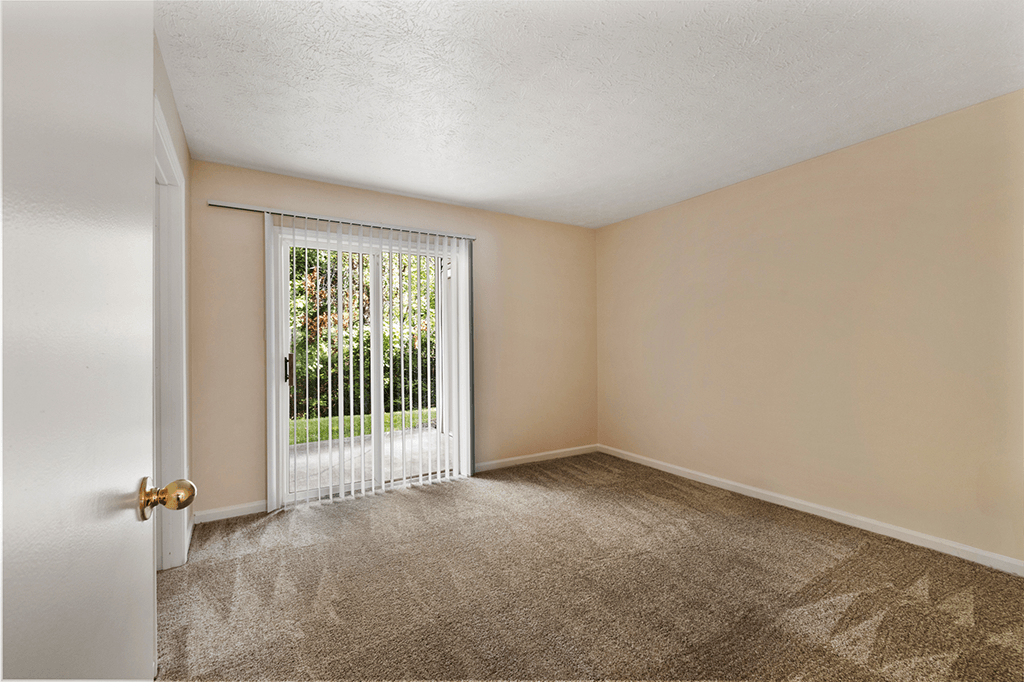 A carpeted living room in a 1-bed, 1-bath apartment at Brownsburg Pointe, featuring natural light from a sliding glass door leading outside.