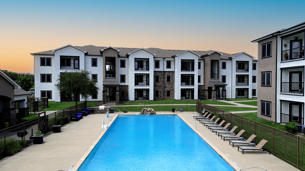 A large swimming pool is surrounded by lounge chairs in front of apartment buildings.
