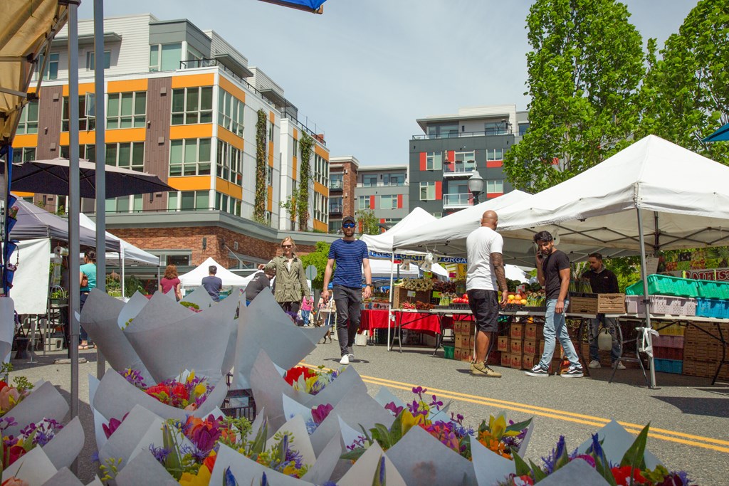 people walking around an outdoor market with stands under umbrellas and selling flowers