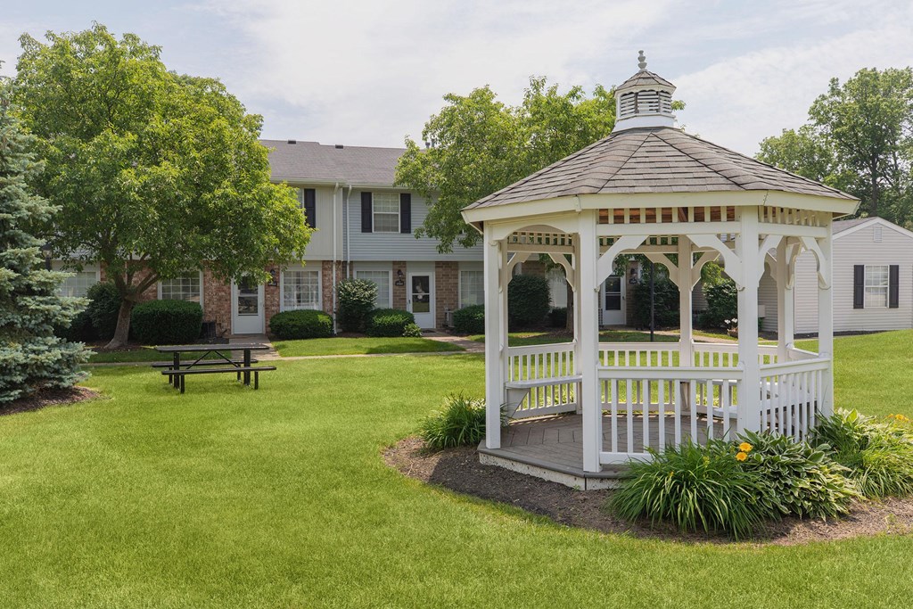 A white gazebo with a black roof is surrounded by a grassy area and a bench.