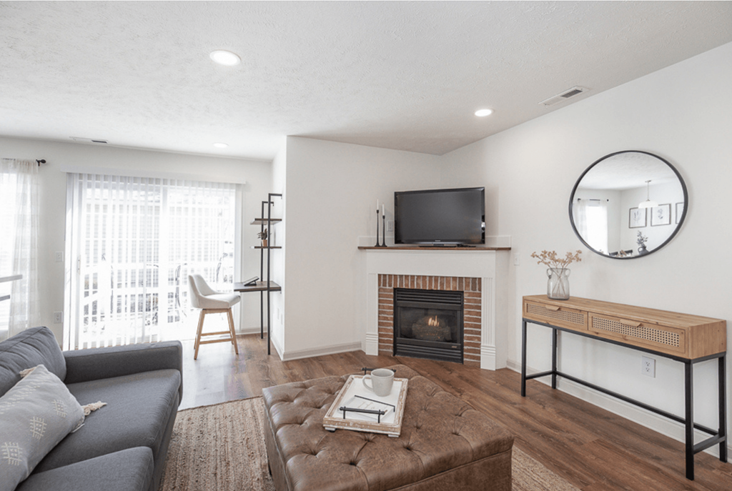 A modern living room with a grey couch, a brown leather ottoman, a fireplace, and a television.
