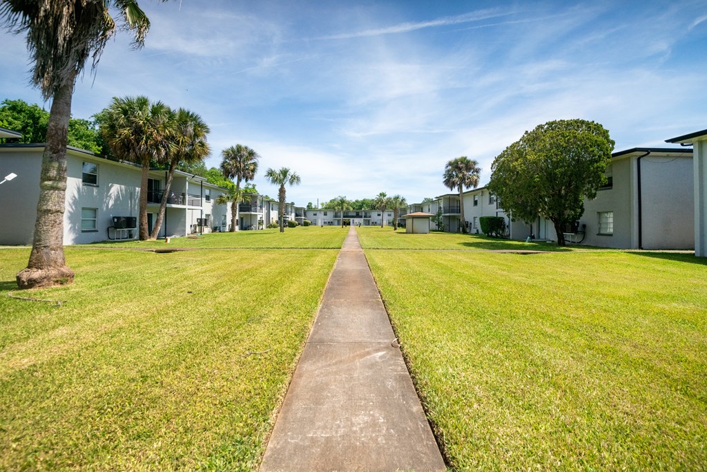 a grassy area with houses on both sides and a sidewalk at Palm Cove Apartments, Daytona Beach, FL
