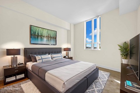 A bedroom with a large bed, a nightstand with a lamp, a painting on the wall, and a window with a view of the sky.