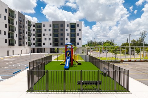 A playground with a slide and a bench is enclosed by a black fence.