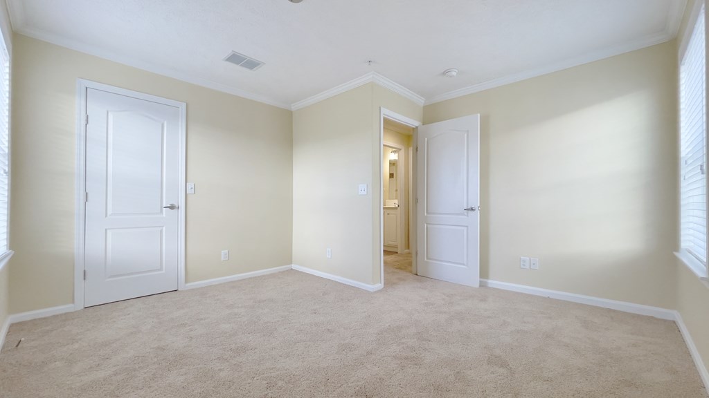 A room with beige carpet and walls, two white doors, and a small window with blinds.