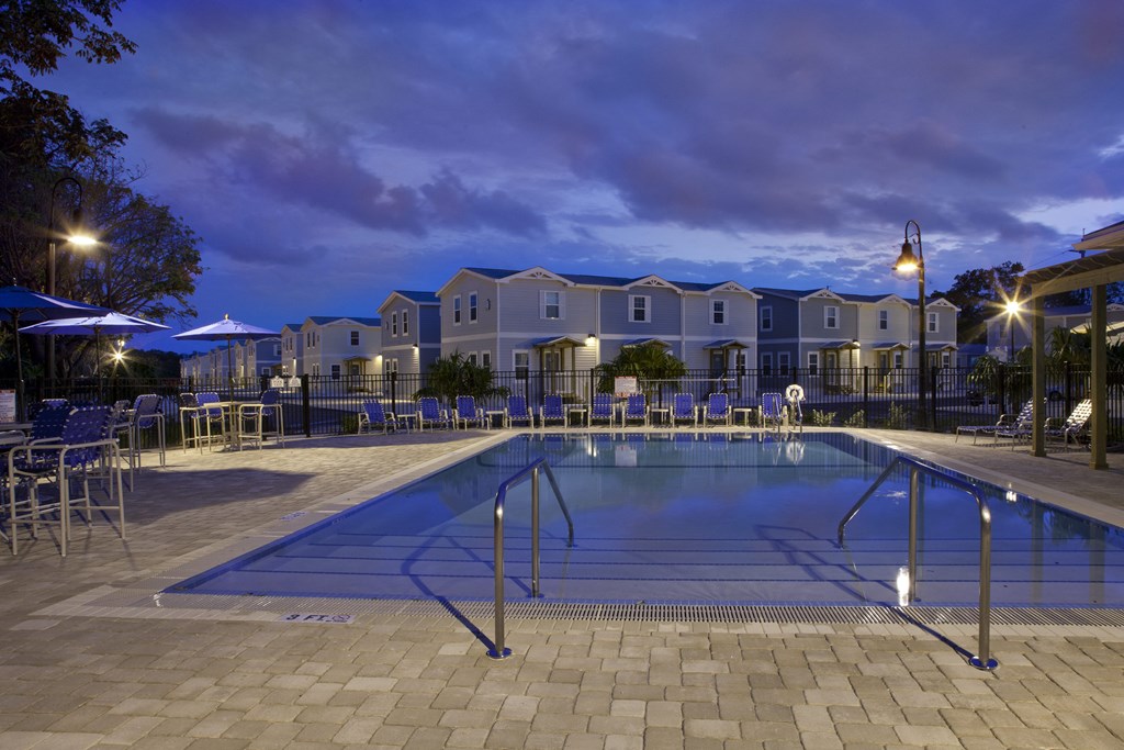 A swimming pool surrounded by a patio and chairs at dusk.