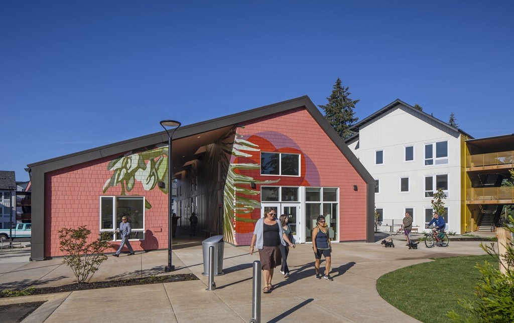 A red building with a white building in the background and people walking in front.