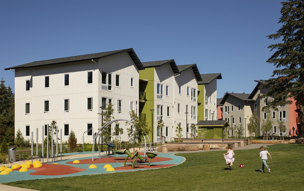 A playground with a colorful play structure is surrounded by a grassy area in front of a white building with a green roof.