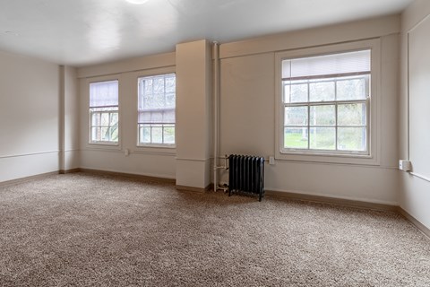 A room with a carpeted floor, two windows, and a radiator.