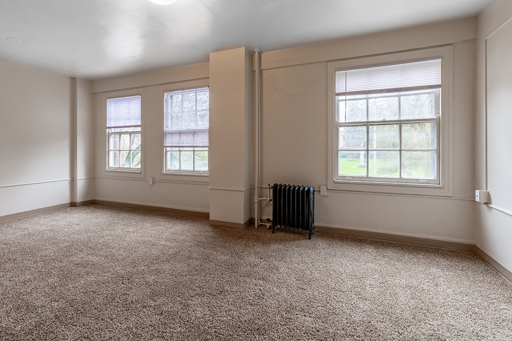 A room with a carpeted floor, two windows, and a radiator.