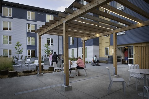A wooden pergola is over a patio with people sitting and a motorcycle parked outside a building.